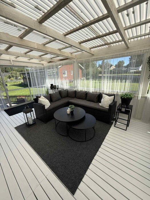 Grå soffa på inglasad altan med svart matta, två svarta runda bord och lantliga detaljer, ljusinsläpp genom transparenta gardiner.