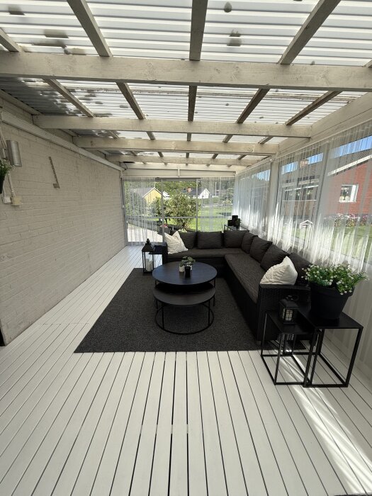 Altan med vitmålade trägolv, en mörk soffa och bord på en matta. Gröna växter och lyktor dekorera hörnet under ett tak av träreglar.