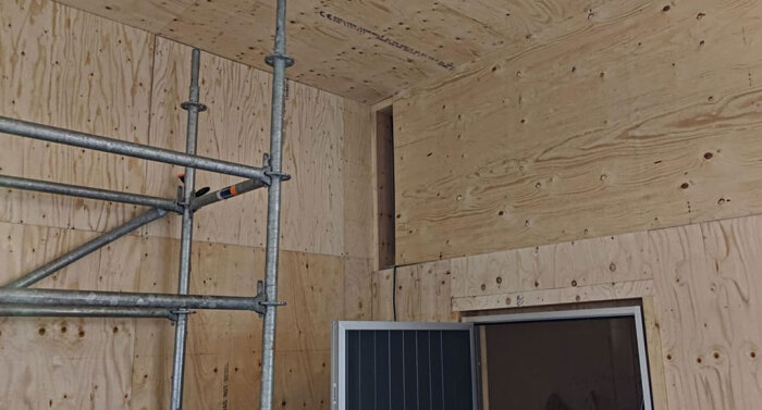Byggarbetsplats med träpanel klädda väggar, ställning och en öppning i väggen. Förberedelse för kabeldragning genom innervägg.