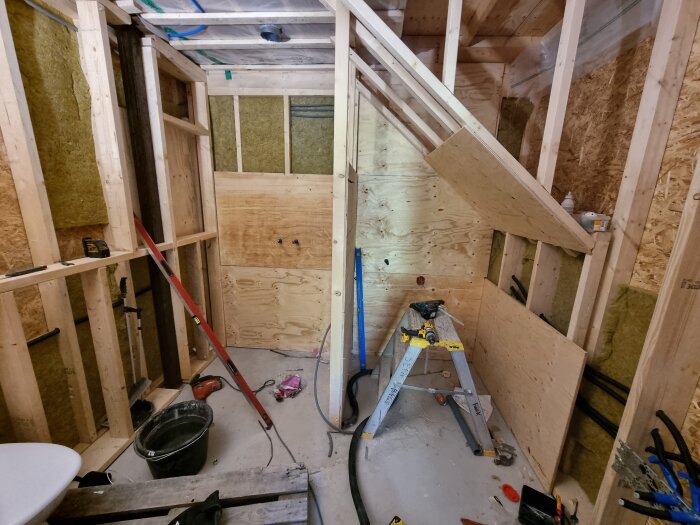 Renoveringsprojekt i ett badrum med träväggar och verktyg som vattenpass, borr och stege. Väggarna är isolerade och träpaneler är delvis monterade.