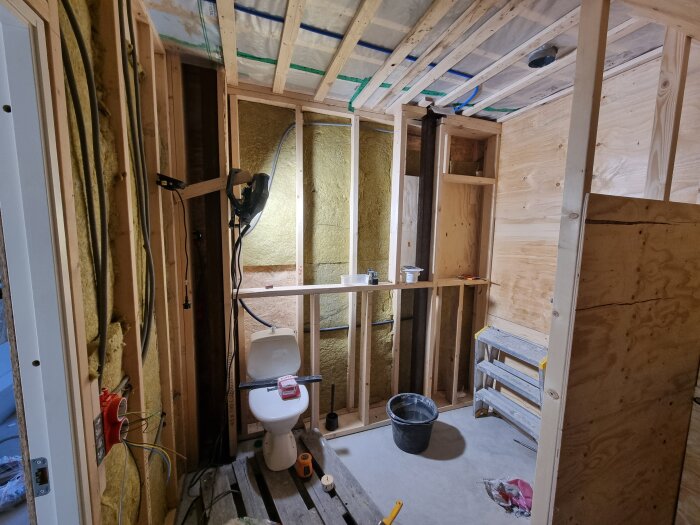 Oinrett badrum under konstruktion med synliga reglar, en arbetslampa, en toalett och en hink på golvet. Stege och plywoodpaneler syns.