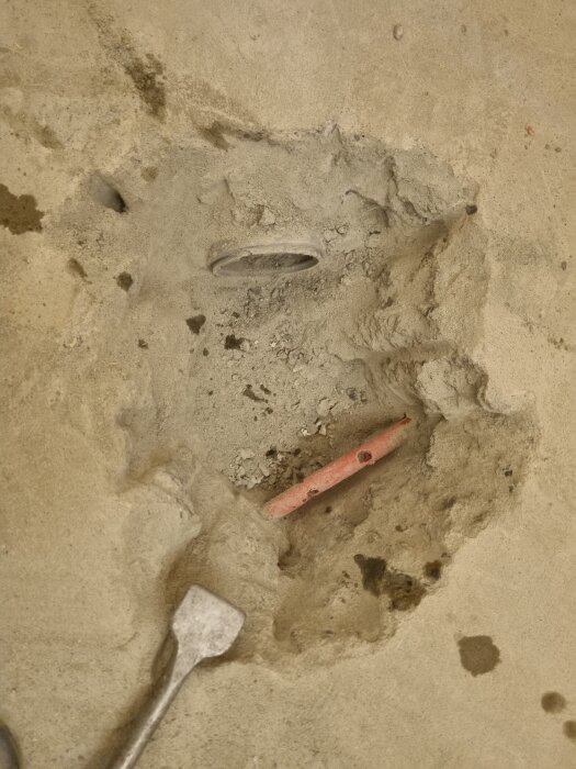 Bild av en delvis uppbruten betonggolv med rör i, där en kakelsättares arbeten påbörjas i ett badrum. En mejsel ligger på golvet.