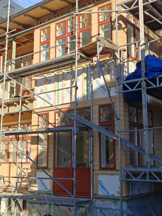 Fasad med röda fönster och dörrar under renovering, omgiven av byggnadsställningar och Glasroc-paneler, saknar fortfarande viss beklädnad.