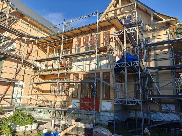 Husfasad under konstruktion med ställningar och färgglada fönsterdetaljer. Byggmaterial och verktyg ligger vid grundarbete.