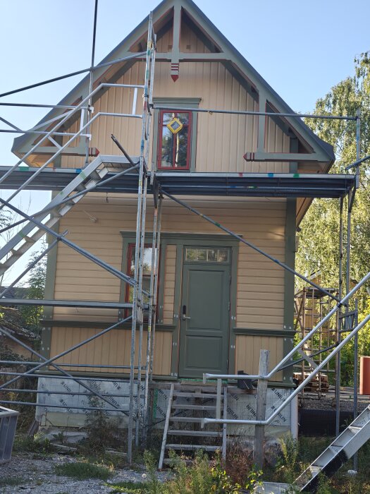 Litet hus med gavel under renovering, omgärdat av byggställningar. Beige och gröna detaljer med påbörjat dekorarbete.