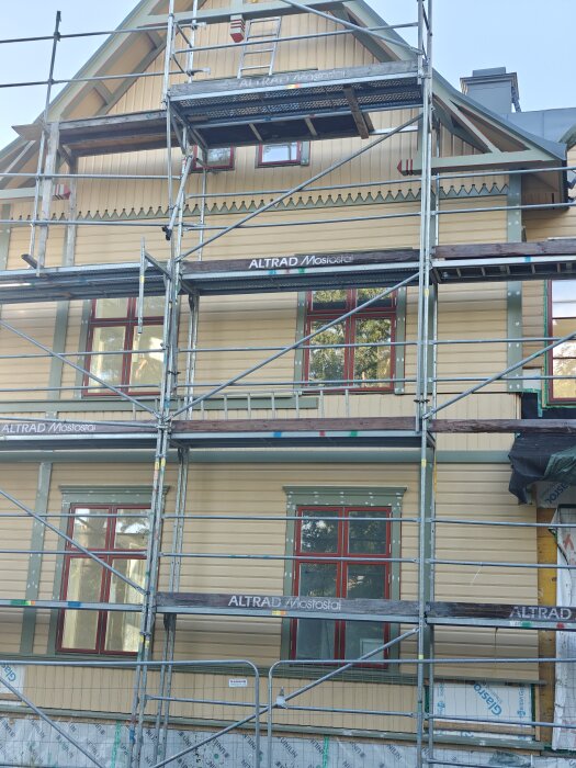 Fasad under konstruktion med byggställning och delvis målad panel samt fönster med röda ramar.
