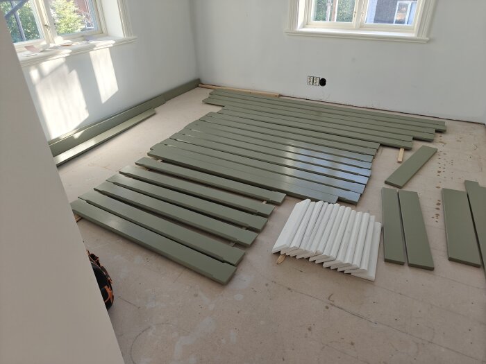 Målade brädor ligger på golvet i ett rum med två fönster. Gröna och vita lister är utspridda, troligen för snickeri eller målning.