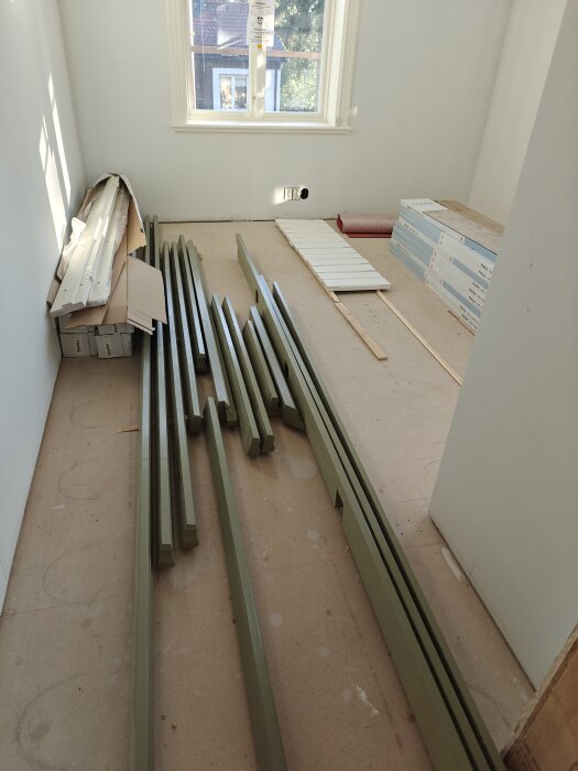 Rum med gröna träbitar och paneler på golvet, bredvid en kartong och en hög med gipsskivor, med ett fönster i bakgrunden.