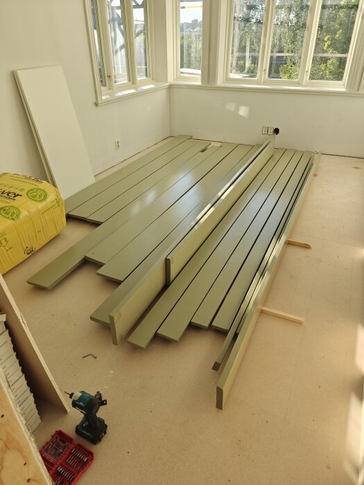 Gröna plankor och byggmaterial ligger på golvet i ett rum med stora fönster. En skruvdragare och låda med verktyg syns bredvid.