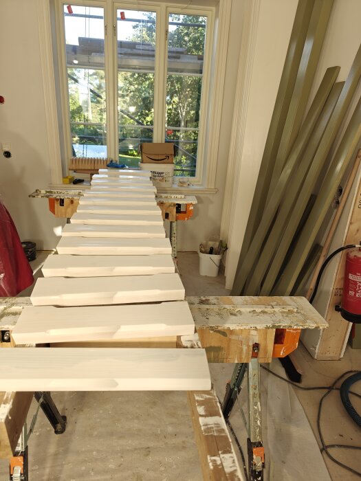 Träpaneler som torkar på en ställning inomhus framför ett fönster, med färgburkar och verktyg i bakgrunden.