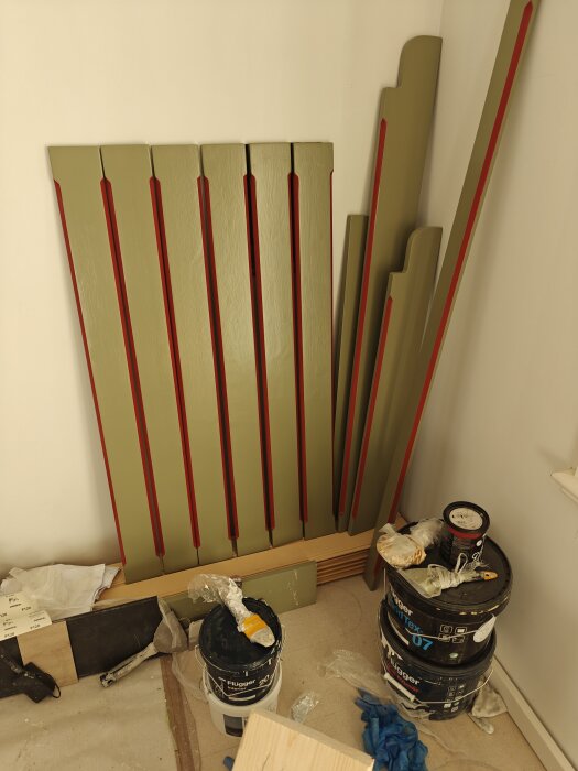 Målade träpaneler och lister i grönt med röda detaljer, omgivna av färgburkar och penslar i ett rum under renovering.