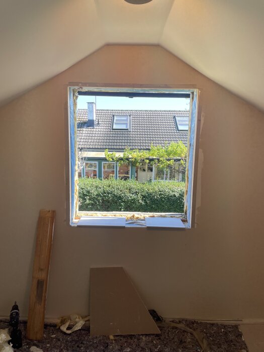 Fönsteröppning i en vägg med fogskum runt kanterna, sett från insidan av ett hus. Tegeltak och växtlighet syns genom öppningen.