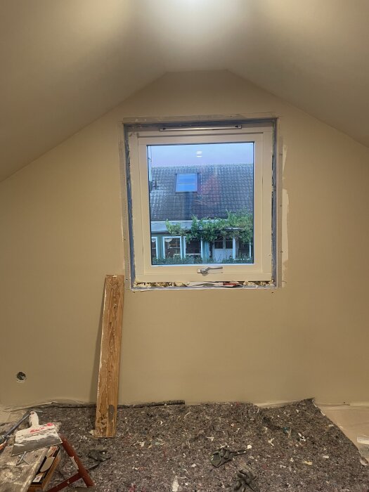 Nytt fönster installerat i ett rum med snedtak, omgiven av ofärdig vägg och byggmaterial på golvet. Trädgårdsscen synlig genom fönstret.
