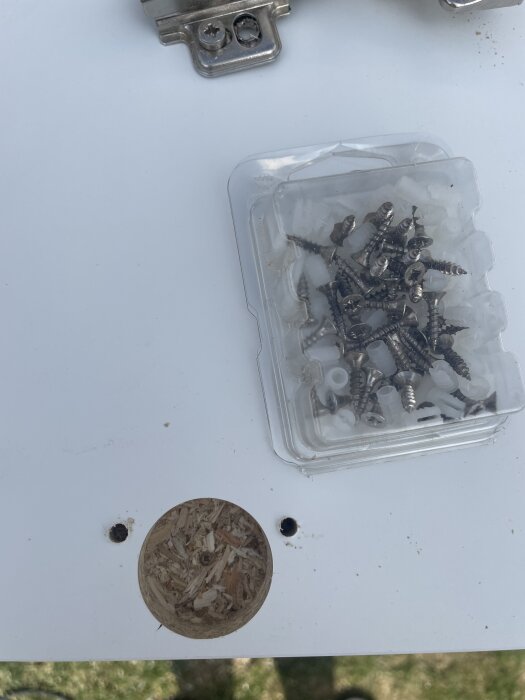 Öppen plastförpackning med skruvar och plugg bredvid borrade hål i en skiva, visar provborrning för gångjärn på IKEA-luckor.