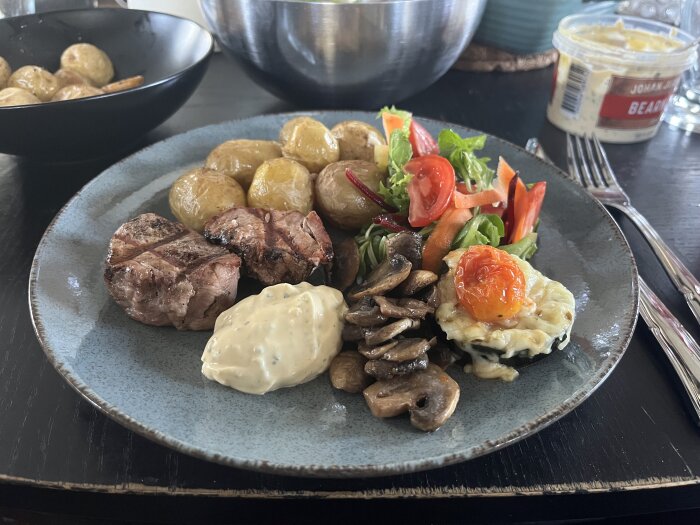 Grillad fläskfilé med ugnstekt potatis, smörstekta champinjoner, sallad, squash med ost och babytomat samt bearnaisesås på en tallrik.