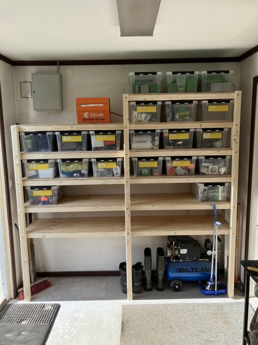 Hylla i garage med plastlådor märkta med verktyg och material, Biltema-kompressor, samt stövlar och sopset vid sidan.