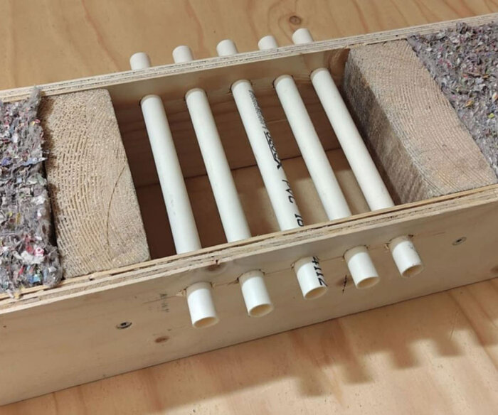 Liten trädummyvägg med sex vita rör insatta mellan träblock. Väggen är gjord av plywood och saknar gips. Rören är placerade horisontellt.