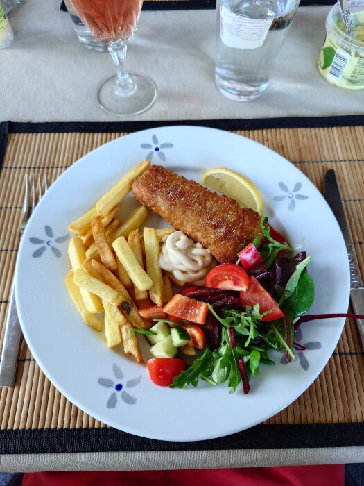 Tallrik med fish and chips, pommes frites, sallad och citronklyfta, plus en klick majonnäs. Vinglas och vattenflaska i bakgrunden.