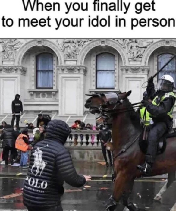 Man med 'Polo'-jacka möter ridande polis i en stadsmiljö, med människor och byggnad i bakgrunden.