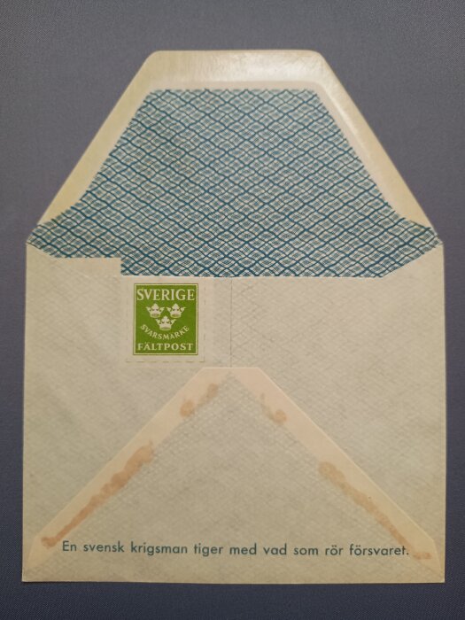 Kuvert med öppet lock, grönt svarsmärke och texten "Fältpost" samt "En svensk krigsman tiger med vad som rör försvaret.