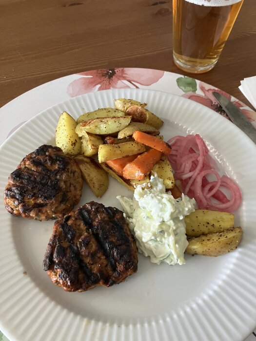 Grillade kycklingfärsbiffar med ugnsrostade potatisar och morötter, picklad rödlök och tzatziki på tallrik, med ett glas öl i bakgrunden.