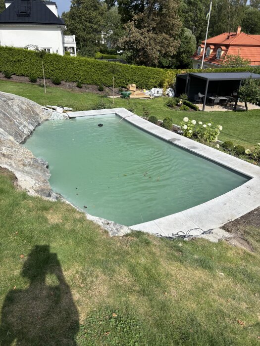 Betongpool med mjölkigt vatten på en bergig bakgård, omgiven av gräs och ett grönskande landskap, med träd och en terrass i bakgrunden.