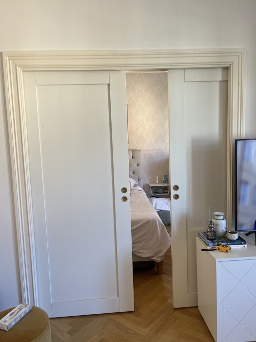 Vita skjutdörrar mellan två rum med glipor i överkant och botten, delvis öppna mot sovrum. Trägolv och vit vägg runt dörröppningen.