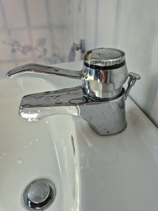 Läckande kromad badrumsarmatur med vatten på toppdelen, sitter monterad på en vit handfat.