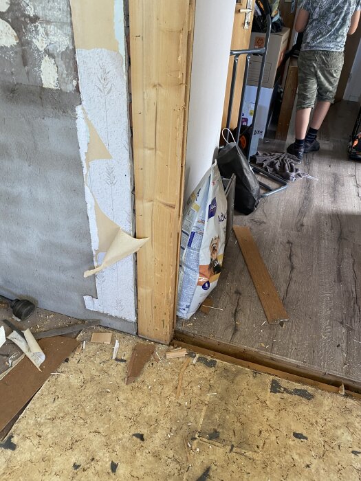 Borttagen dörrkarm i renoveringsprojekt, exponerat timmer mot murstock, golvdelar och byggmaterial synliga, pågående arbete med öppning mot kök.