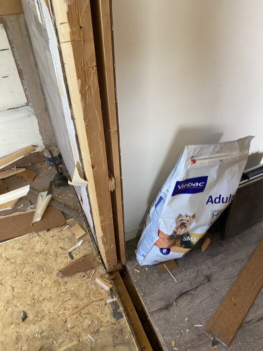 Timmer bredvid murstock, borttagen dörrkarm, spånskivor på golvet, påse hundmat lutad mot väggen, renoveringsstök i hall.