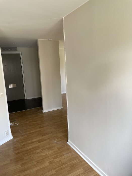 Målad vägg med ojämnheter i nyrenoverad lägenhet, golv med ljus träfinish och dörr i bakgrunden.