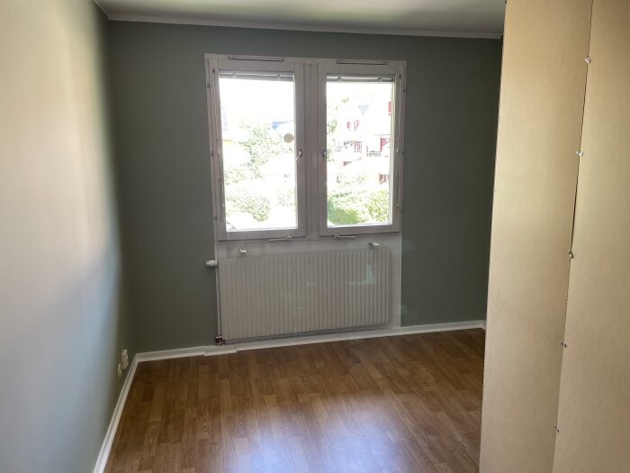 Tomt rum med gröna väggar, brunt golv och vitmålat fönster. Element under fönstret; bilden visar underbehandlingens ojämnheter.