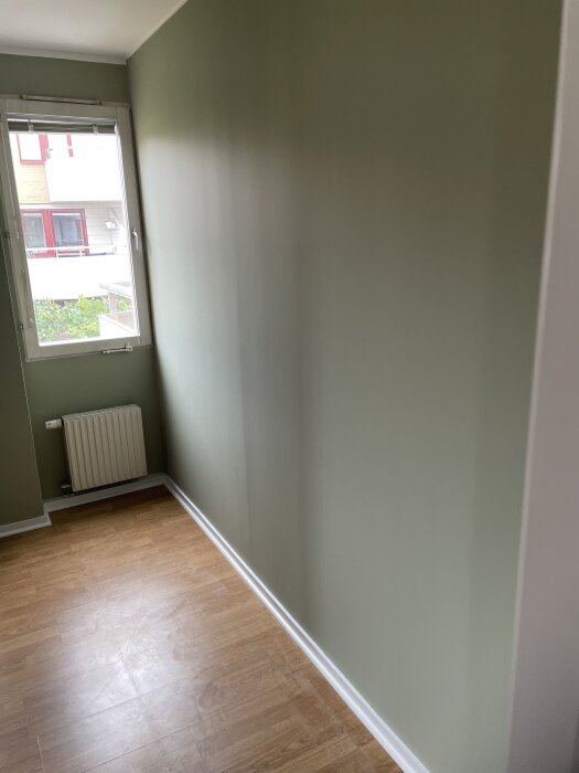 Grönmålad vägg med vit list och brunt trägolv i rum med fönster och element. Slät yta men ev. tecken på tidigare ojämnheter.