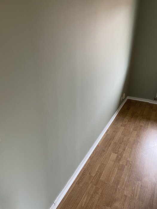 Nymålad vägg med ljusgrön färg där ojämnheter syns, vit list längs golvet, parkettgolv av trä.