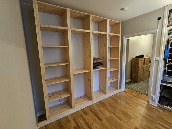 Hemmagjord garderobsstomme av trä utan dörrar, byggd av 18mm skivor och 21mm plankor, med öppna hyllor delvis fyllda med grå och bruna kuddar.