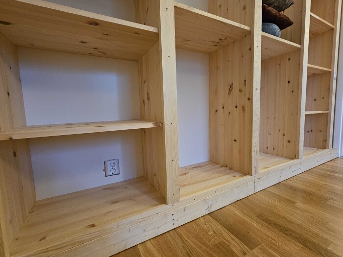 Trähylla byggd av 18mm skivor och 21mm plankor utan dörrar, med öppna fack och en eluttag på väggen bakom.