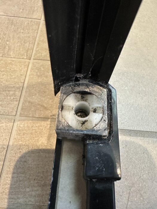 Svart duschdörr med trasig vit plastdel i botten, delvis smutsig och på ett klinkergolv.