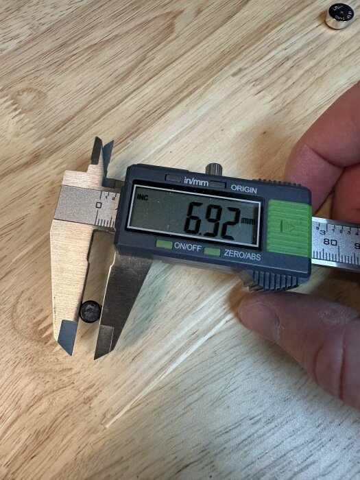 Digitalt skjutmått visar diameter 6,92 mm på en liten rund plastdel, liggande på ett träbord.