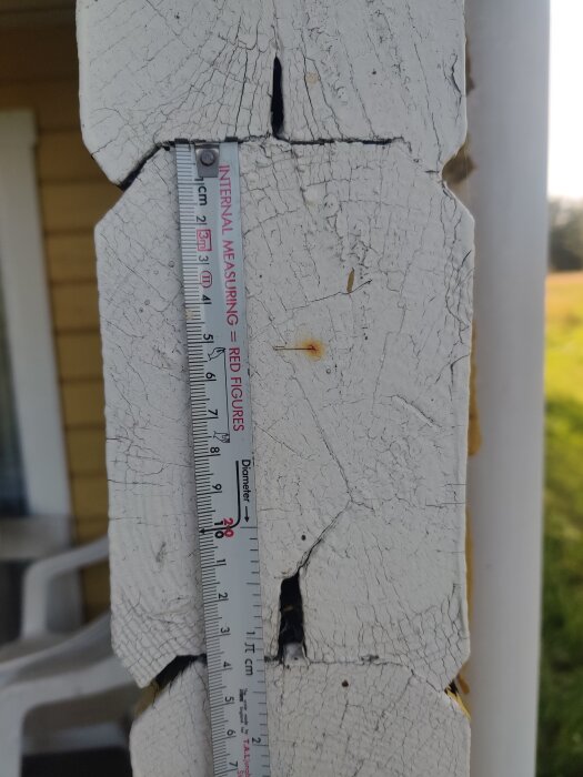 Träprofil med måttband, vitmålad och sprucken, sökes för renovering av 60-tals husbyggsats; står mot gul byggnadsvägg.