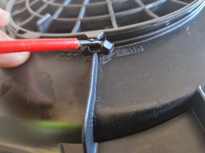 Röd skruvmejsel bänder upp låsflärp på svart plastkonstruktion, del av motor.