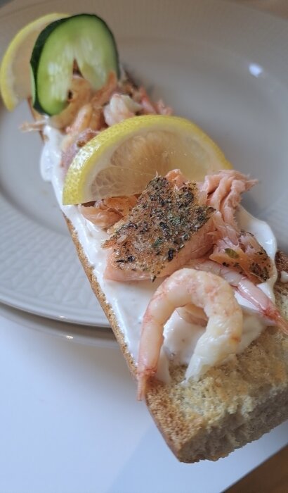 Rostad baguette med aioli, örtkryddad lax, rökta räkor, kräftstjärtar, citron- och gurkskiva, på en tallrik.