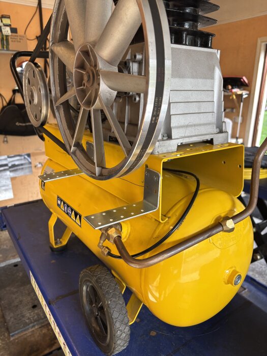 Gul kompressor med ny oljekylare och kopplingar, placerad på ett arbetsbord i ett garage.