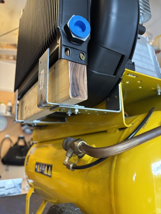 Ombbyggd kompressor med monterad oljekylare och kopplingar, gult tankhölje och kopparrör. Trädelar används för stabilisering.