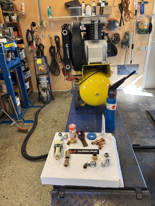 Kompressor med gul tank i verkstad, omgiven av verktyg och komponenter som kopplingar och en oljekylare, på ett bord framför.