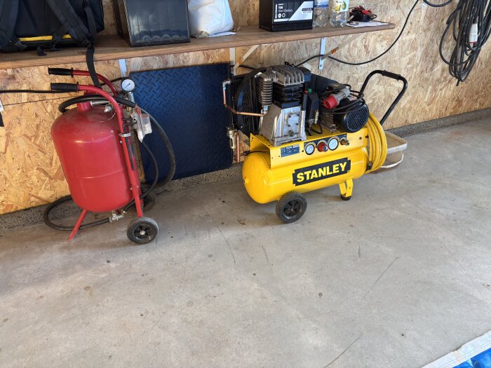 Gul Stanley-kompressor vid en verkstadshylla bredvid en röd tryckbehållare på hjul, redo för att testa torrare luftproduktion.