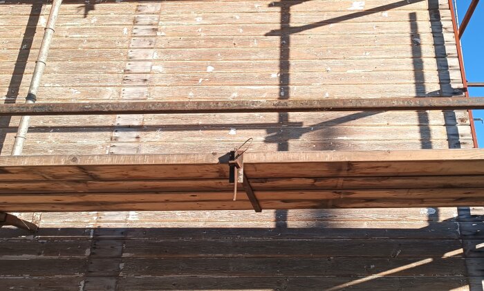 Byggnadsställning mot en träfasad med avskrapad färg i solsken.