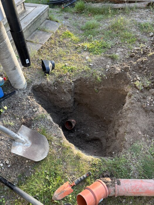 Grävd grop bredvid ett hus med synliga dagvattenrör och verktyg. Problem med vattenansamling i rör som diskuteras för lösning.