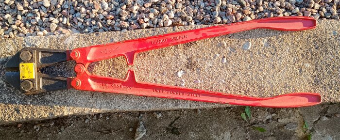 Röd bultsax från Bahco, liggande på en stenig yta. Verktyget har en robust design med skarpa skärverktyg.