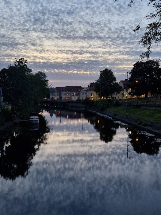 Kvällsbild av en stilla flod med en småbåt, hus och träd reflekterade i vattnet, molnig himmel ovanför, gatlyktor lyser svagt.