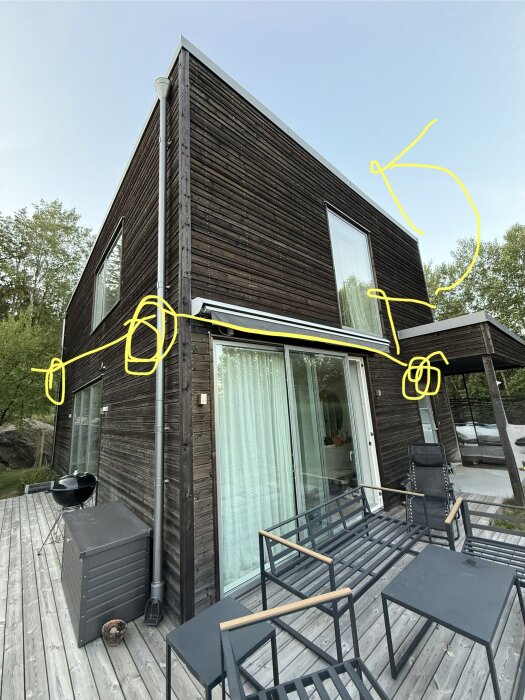 Fasad av ett modernt hus med träpanel där kamerainstallationer planeras, markerade med gula cirklar och pilar. Träveranda med utemöbler.
